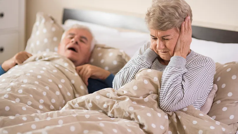 Un'immagine di una coppia di anziani a letto, dove il marito russa e tiene sveglia la moglie.