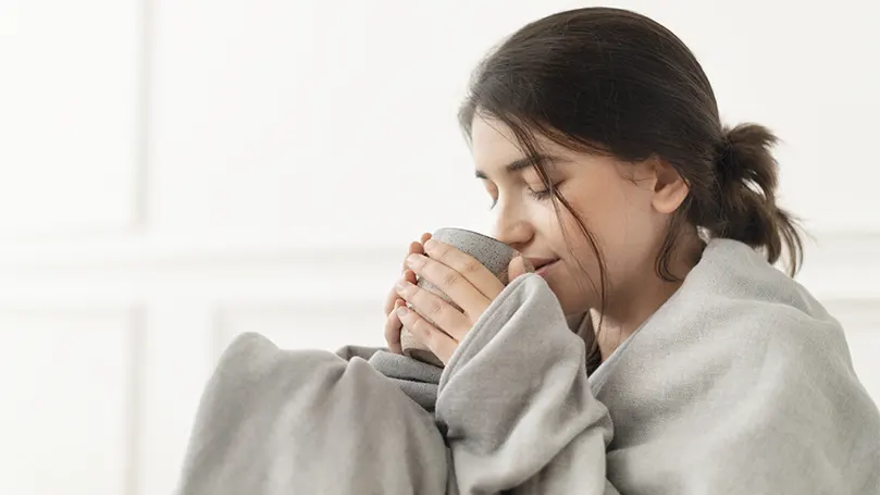 Un'immagine di una donna che beve il tè avvolta in una coperta ponderata per la stimolazione della pressione profonda.