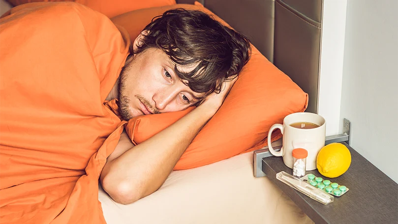 Un uomo sdraiato su un letto malato e coperto da una coperta arancione.