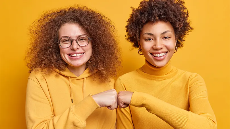 Un'immagine di due donne con maglioni arancioni che si stringono i pugni e si sostengono a vicenda.